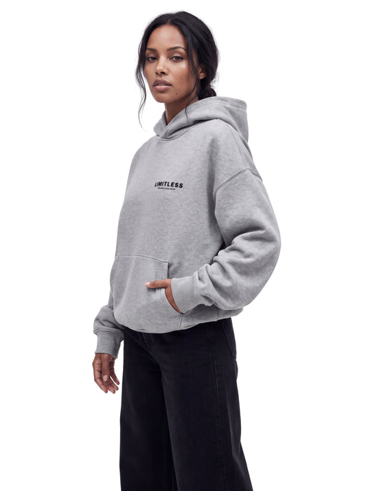 Woman in oversized heather grey LIMITLESS fleece hoodie and baggy black jeans, three-quarter view with hand in pocket